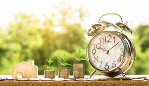 An alarm clock, three piles of coins, and a small wooden model house symbolizing the Dammam real estate market.